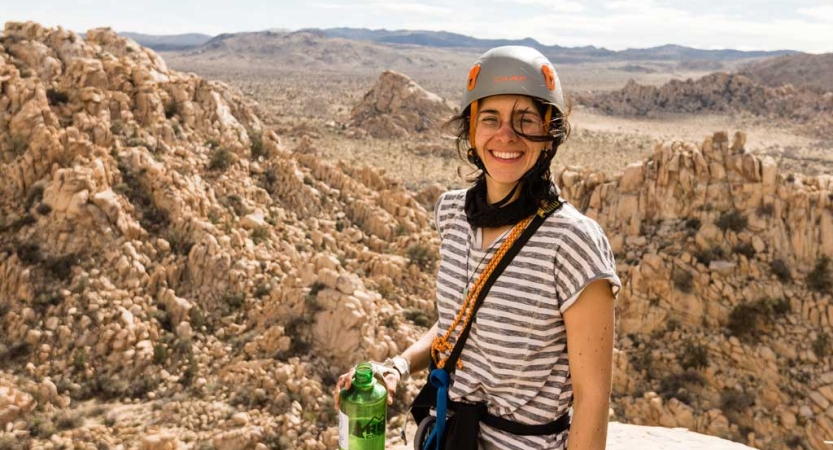 A person wearing a helmet stands and smiles on high ground, far above a vast rocky landscape below. 